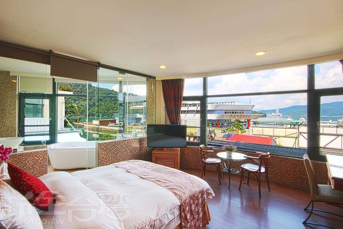 A hotel room with a bed, a TV, a small table with chairs, and a large window offering a view of a body of water and buildings.