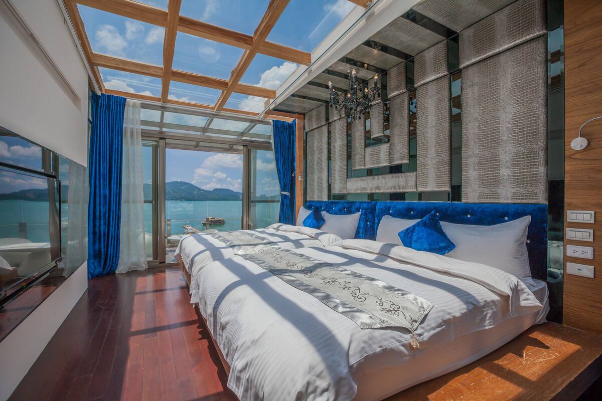 A bedroom with a large bed, a glass wall, and a view of the sea.