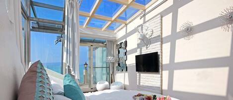 A bedroom with a large skylight, a bed with multiple pillows, a mounted TV, and a view of the sea.