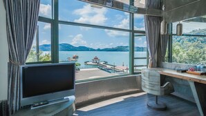 A room with a large window offering a view of a lake and mountains, a TV on a stand, and a desk with a chair. - Shuian Lakeside Hotel (Yuchi)
