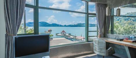 A room with a large window offering a view of a lake and mountains, a TV on a stand, and a desk with a chair.