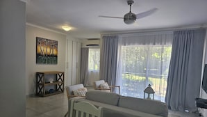 Un salon moderne avec un canapé, des fauteuils, une console en bois, un tableau et une vue sur un jardin grâce à de grandes fenêtres.
