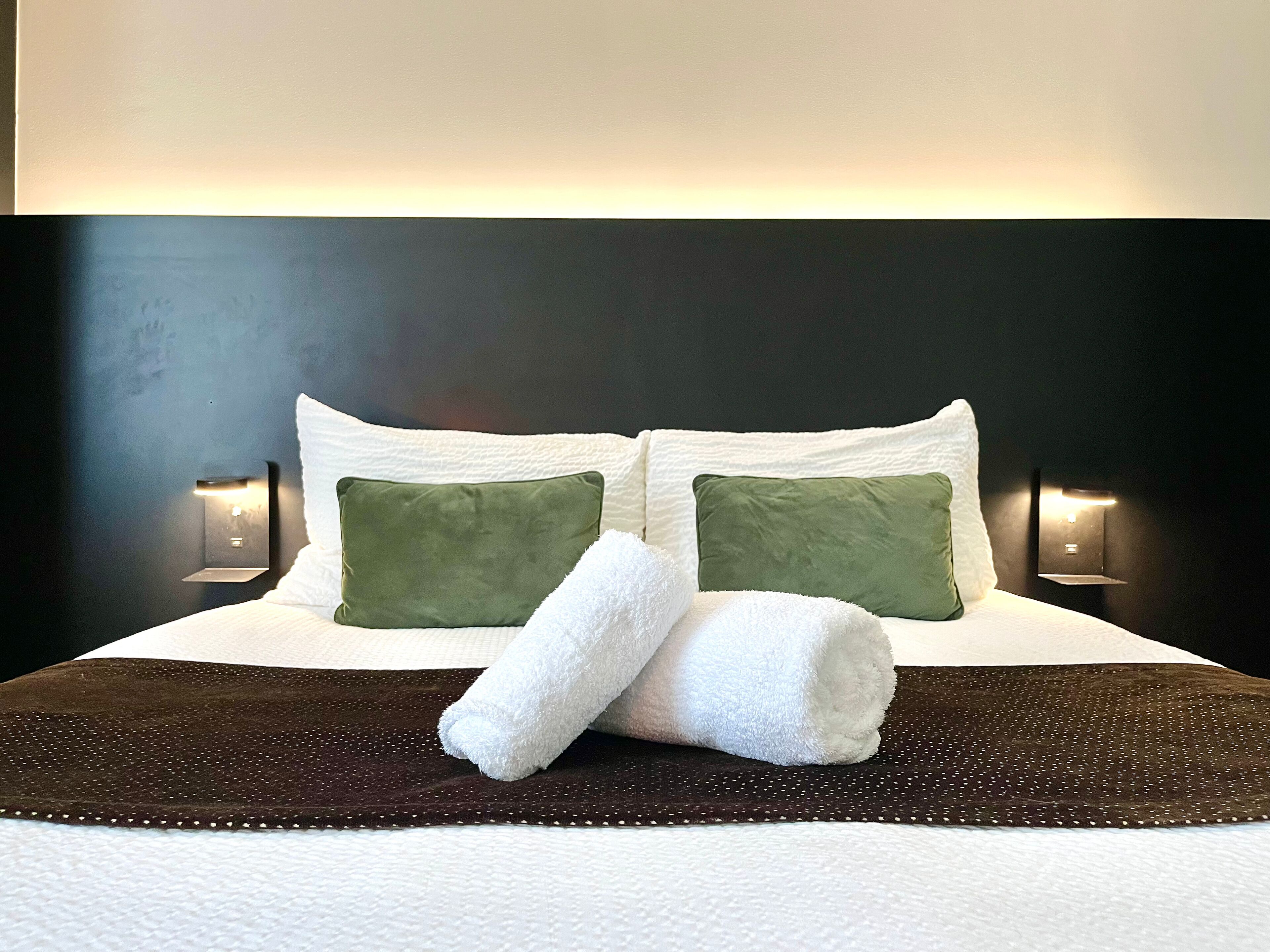 Una cama bien tendida con dos almohadas, una toalla blanca enrollada y una colcha marrón oscuro.