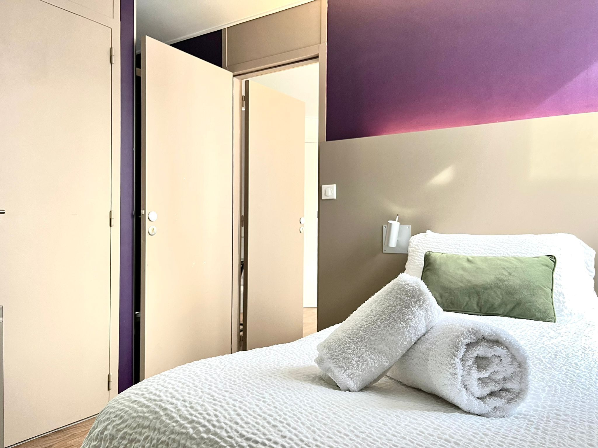 A hotel room with a bed, a green pillow, and rolled towels.