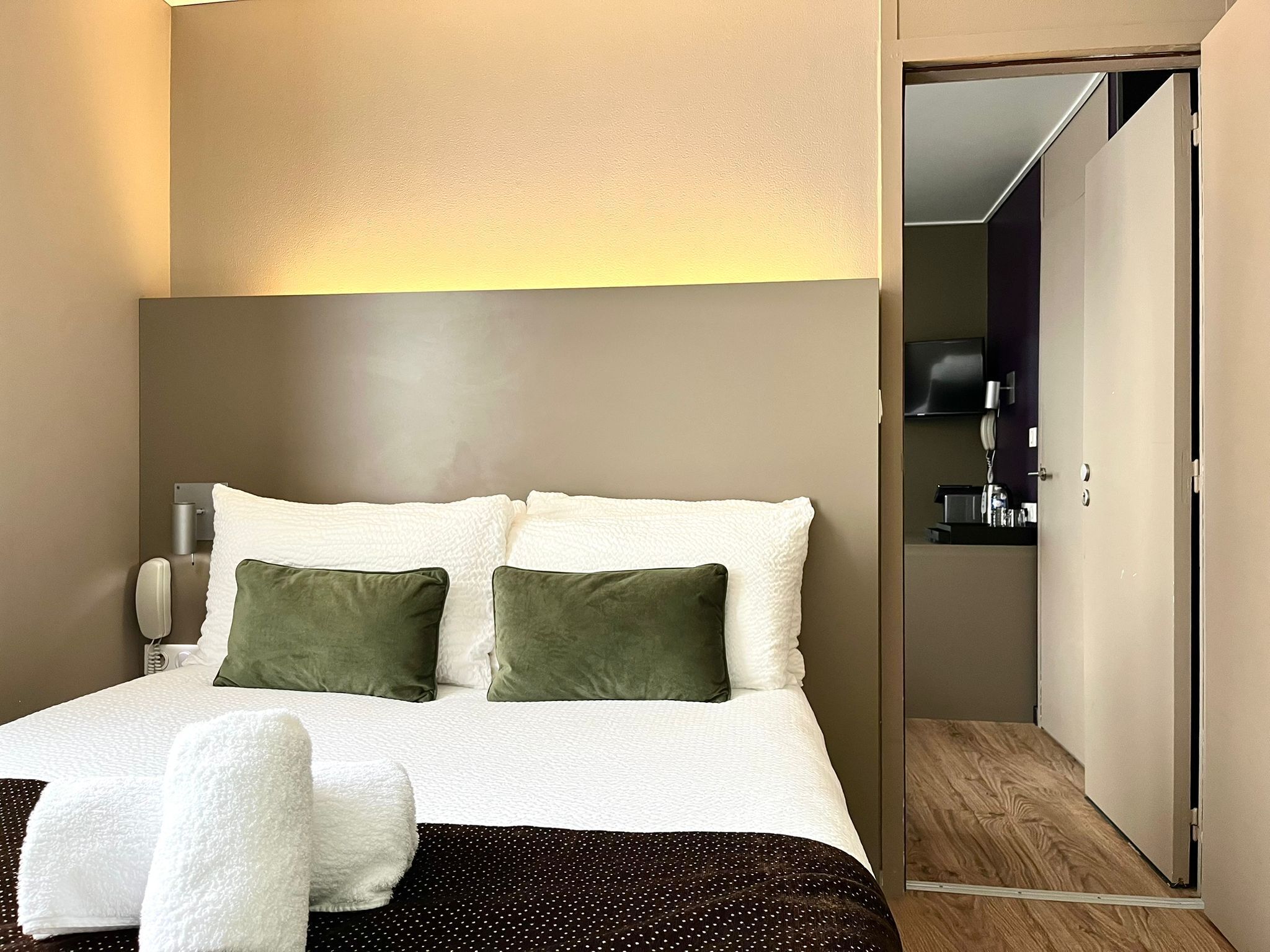 A hotel room with a bed, two pillows, a bedside table, a lamp, and a wooden floor.
