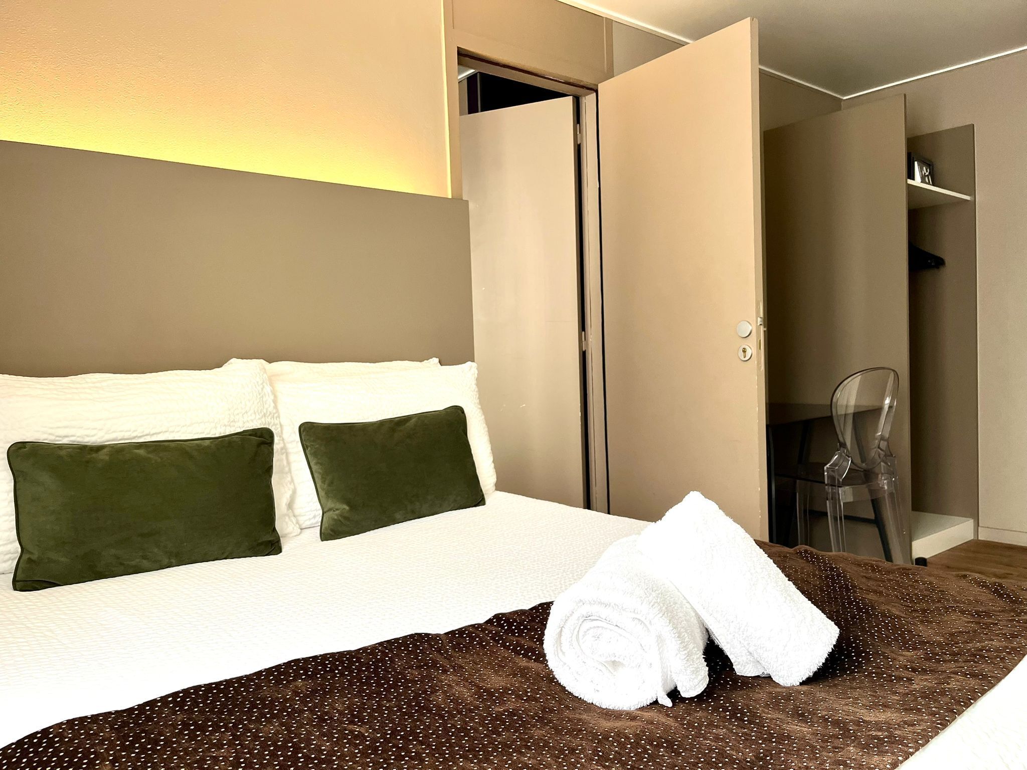 A hotel room with a bed, two pillows, a rolled white towel, and a brown blanket.