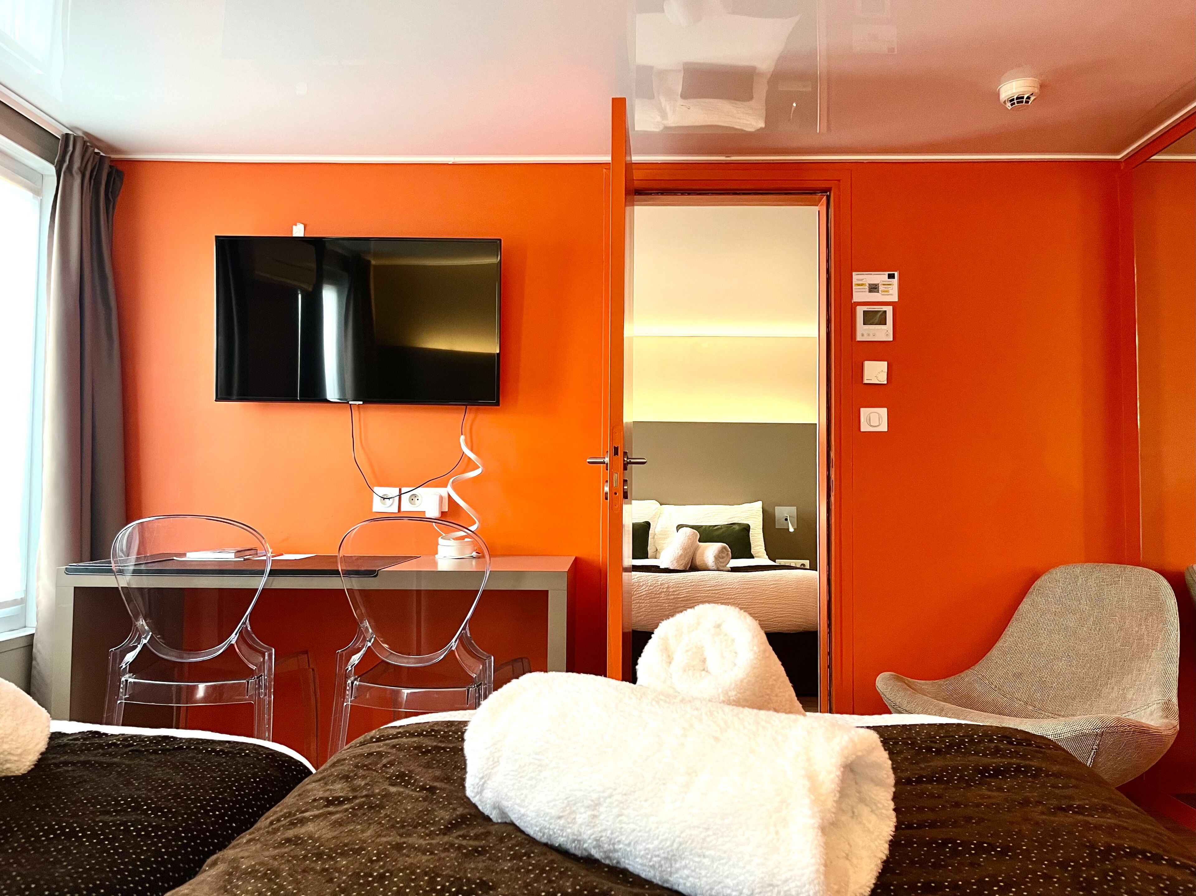 Een hotelkamer met een oranje wand, een televisie boven een bureau, twee stoelen, een bed met witte handdoeken en een deur naar een andere kamer.