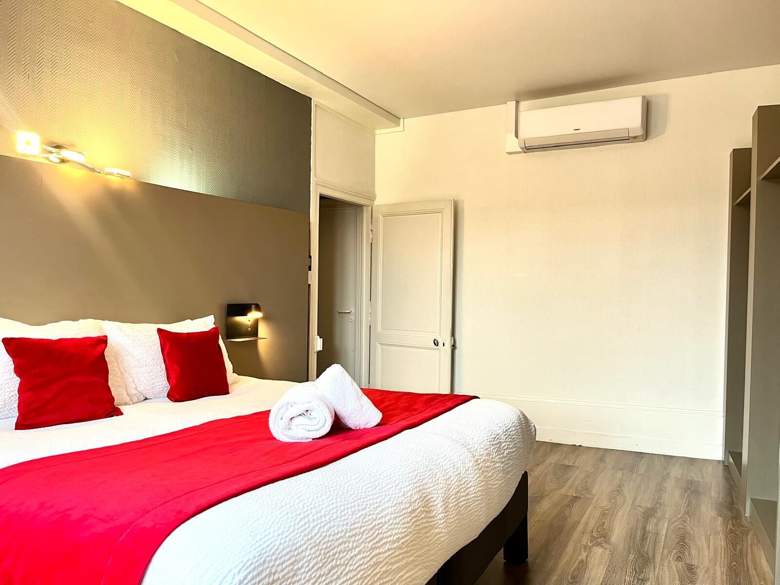 A hotel room with a bed, red and white bedding, a wall-mounted air conditioner, and a door leading to another room.