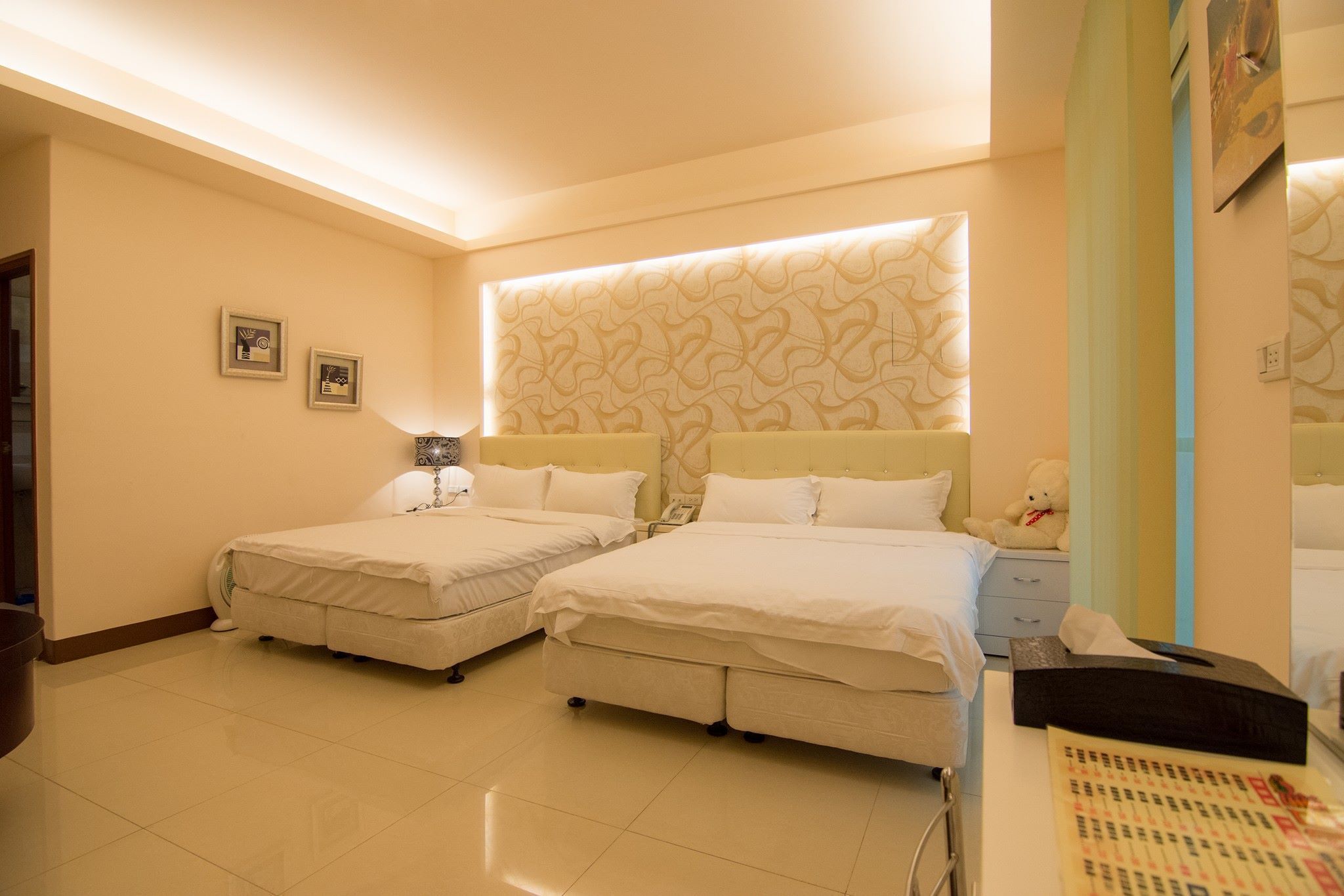 A hotel room with two beds, a bedside table with a lamp, a teddy bear, and a wall with a decorative pattern.