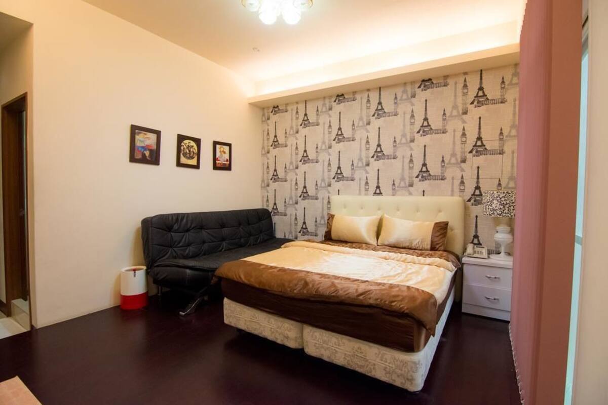 A bedroom with a bed, a black sofa, a nightstand, and a wall with Eiffel Tower wallpaper.