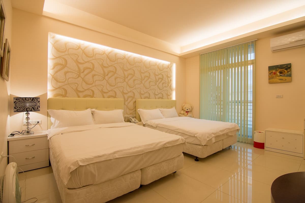 A hotel room with two beds, a bedside table with a lamp, a wall-mounted air conditioner, and a window with blinds.