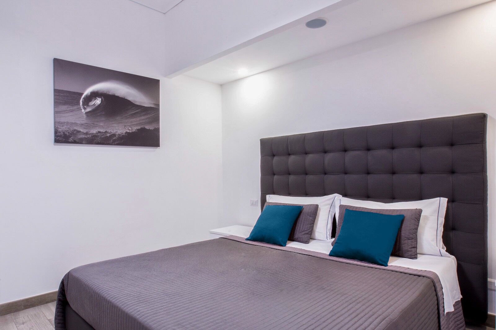 A modern bedroom with a large bed, a tufted headboard, and a framed picture of a wave on the wall.