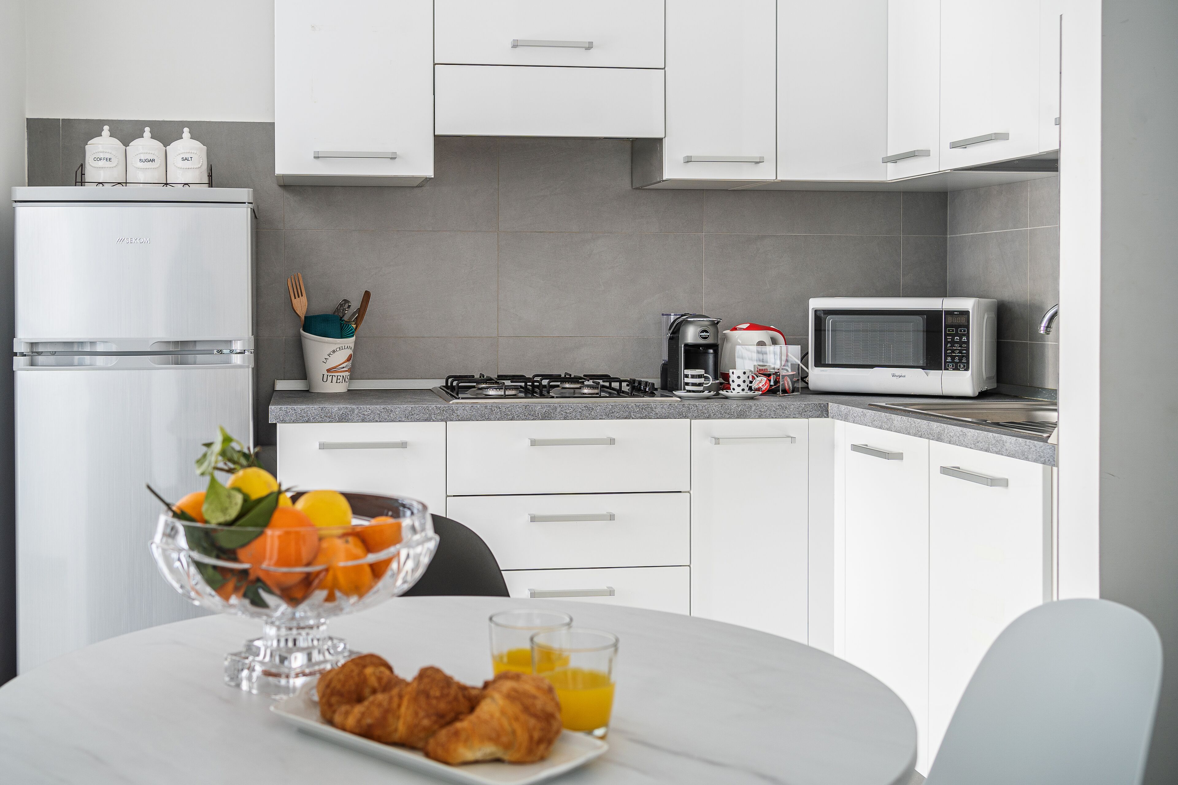 Una cocina moderna con armarios blancos, un refrigerador de acero inoxidable, un microondas y una estufa a gas. Hay una disposición para el desayuno en la encimera con un frutero, croissants y vasos de jugo de naranja.