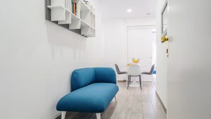 Een moderne, minimalistisch ingerichte kamer met een blauwe bank, witte open planken en een eethoek met een tafel en stoelen.