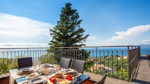 Une table à manger dressée pour quatre personnes, avec des assiettes, des verres et une variété de plats, offrant une vue pittoresque sur la mer et un arbre.