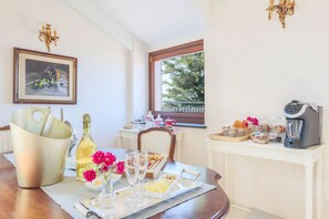 A breakfast table set with wine, flowers, and pastries, a coffee maker, and a view of trees through a window.