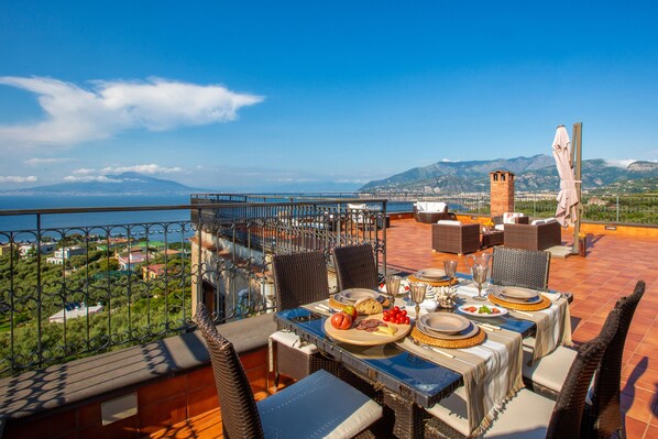 A terrace with a table set for dining, overlooking a coastal landscape with mountains in the distance.