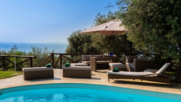 Un espace aménagé au bord de la piscine, avec des chaises longues et une table placée sous un parasol, offrant une vue sur une étendue d’eau et des montagnes lointaines.