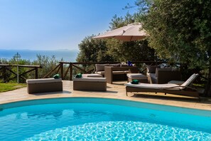 A poolside area with lounge chairs and a table under an umbrella, overlooking a body of water and distant mountains.