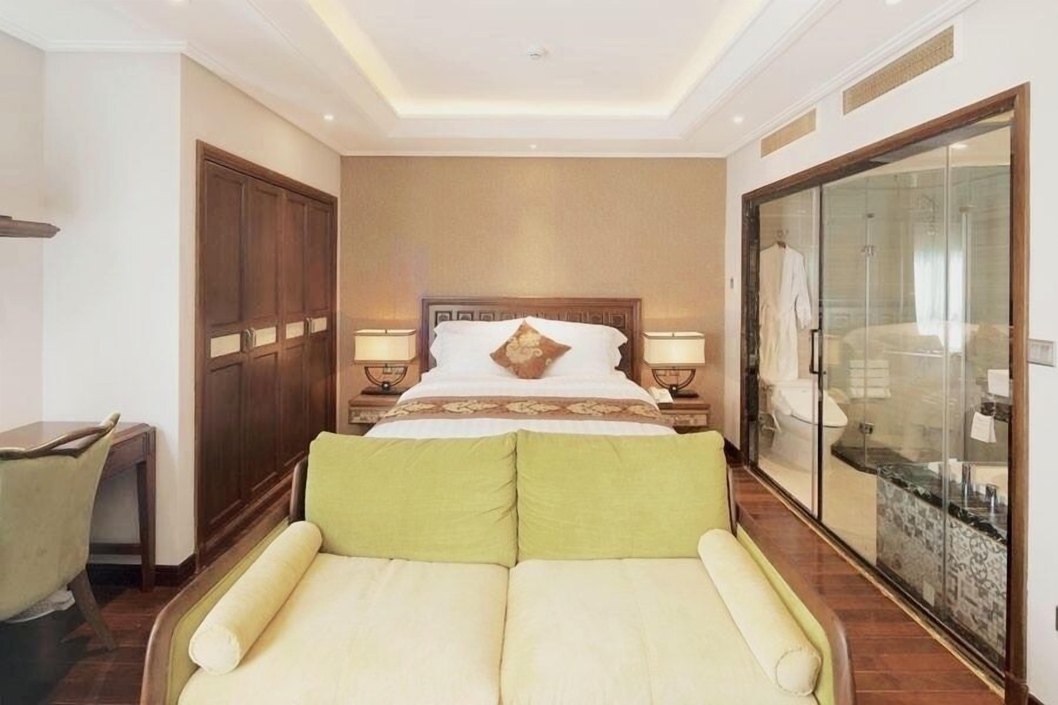 A hotel room with a large bed, bedside tables with lamps, a wardrobe, a seating area with cushions, and a glass shower enclosure.