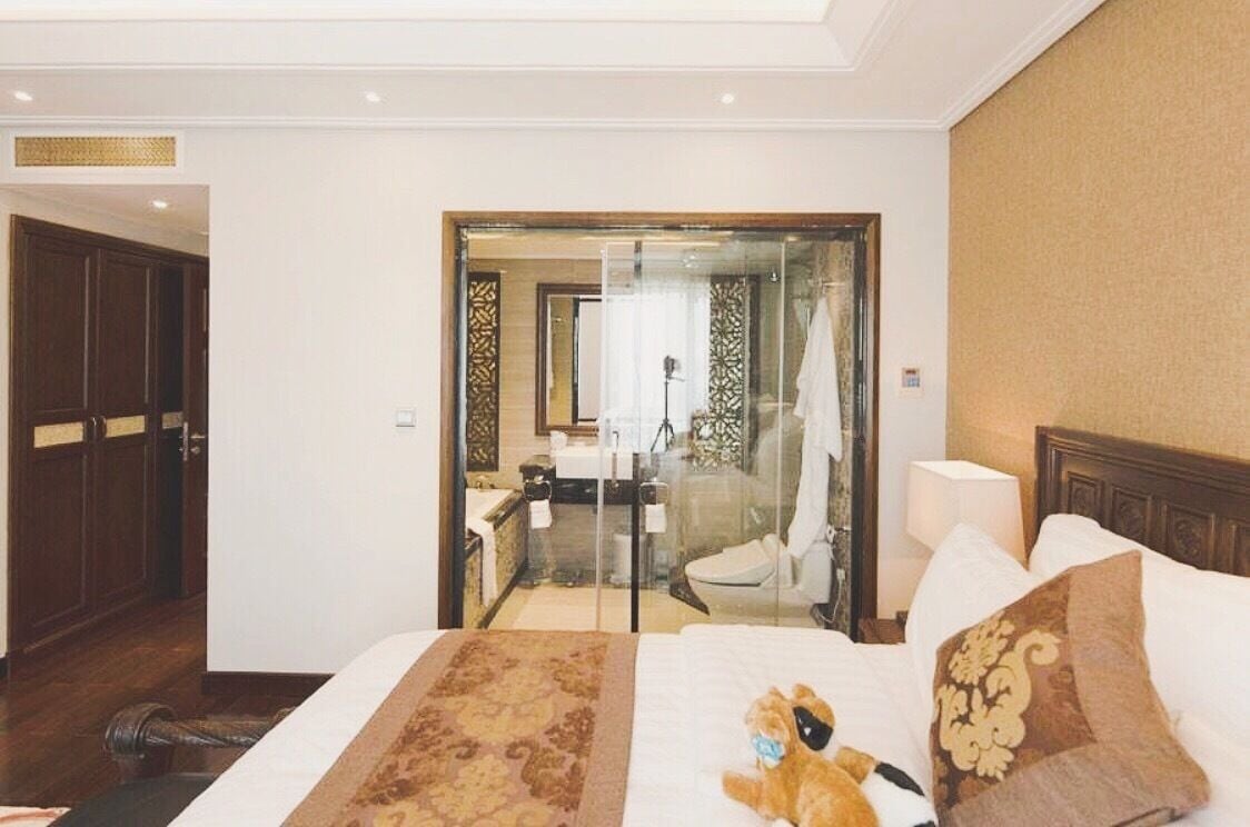 A hotel room with a large bed, a plush teddy bear, a nightstand, and a bathroom visible through a glass door.