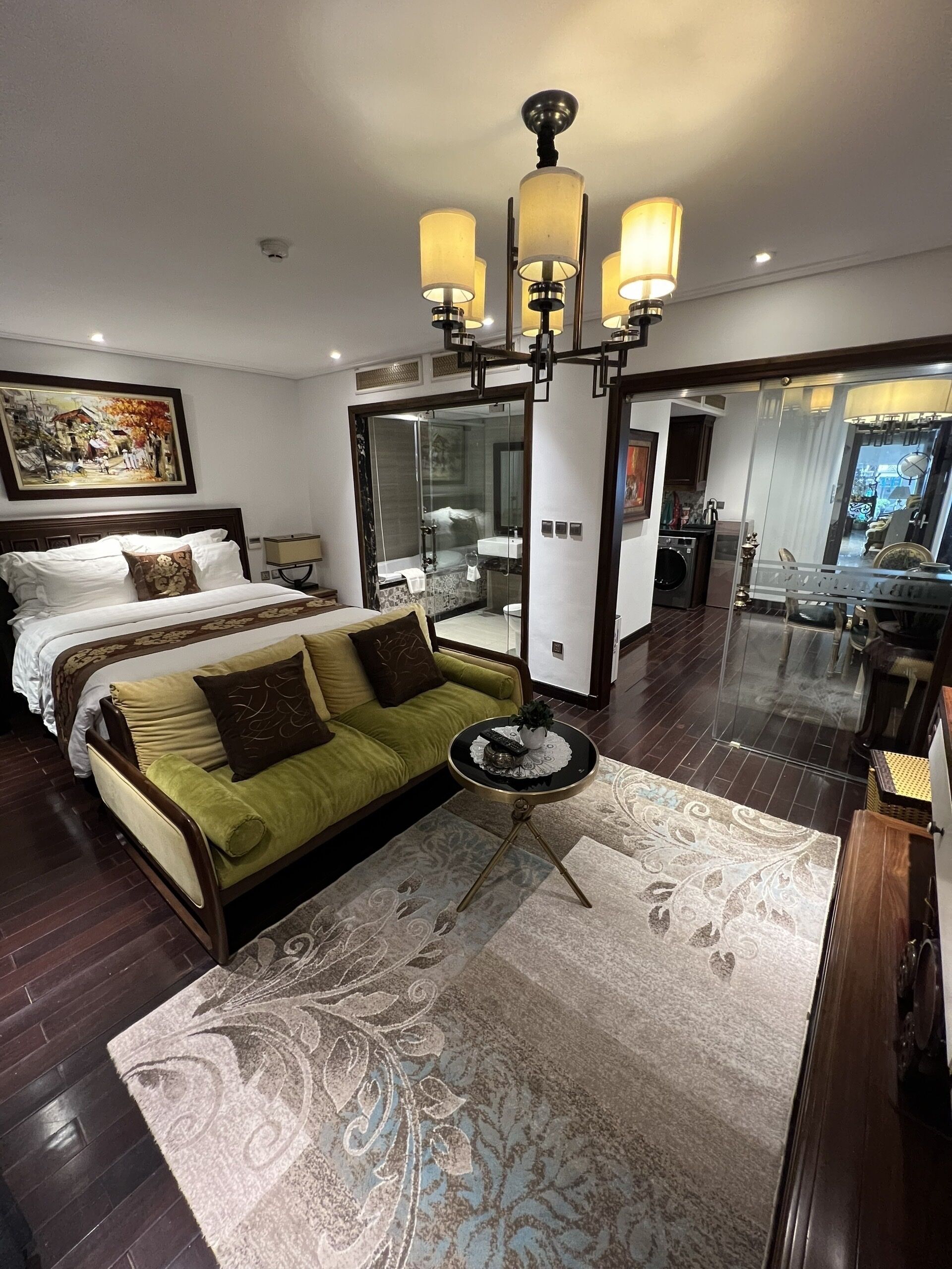 A modern hotel room with a large bed, a green sofa, a round coffee table, and a glass-enclosed shower area.