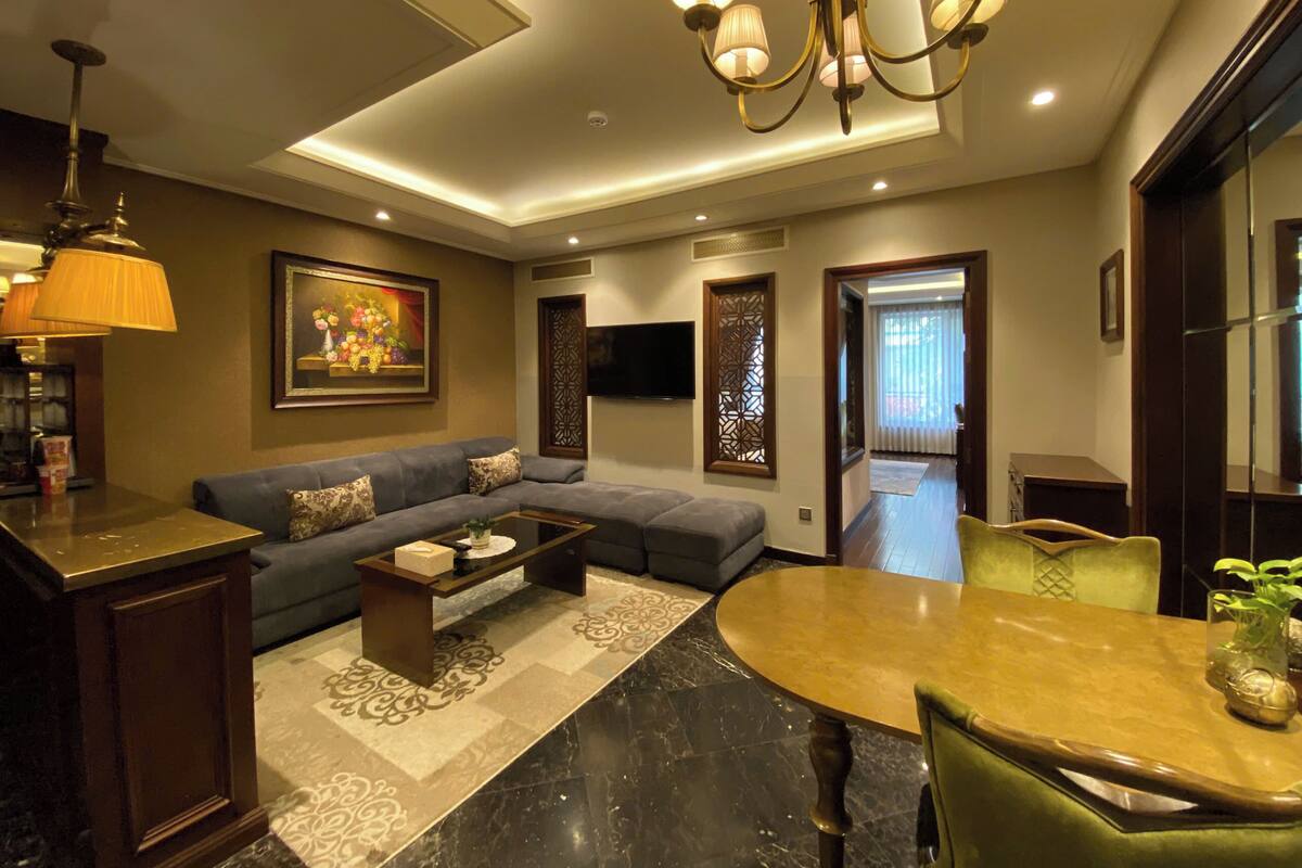 A modern living room with a sofa, coffee table, and dining table. There is a chandelier, a painting on the wall, and a TV mounted above the sofa.