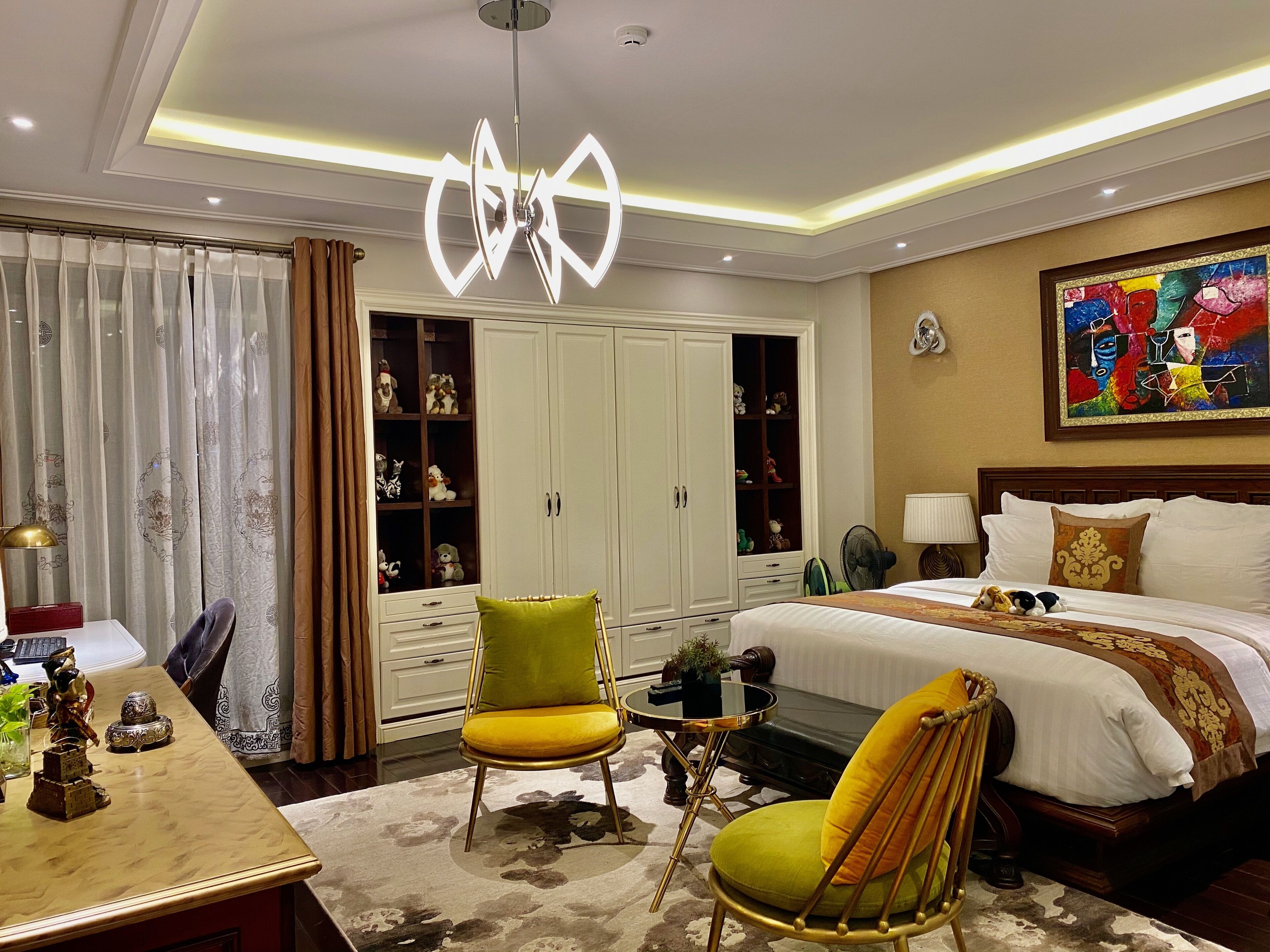 A bedroom with a bed, two chairs, a desk, a wardrobe, and a ceiling light fixture.