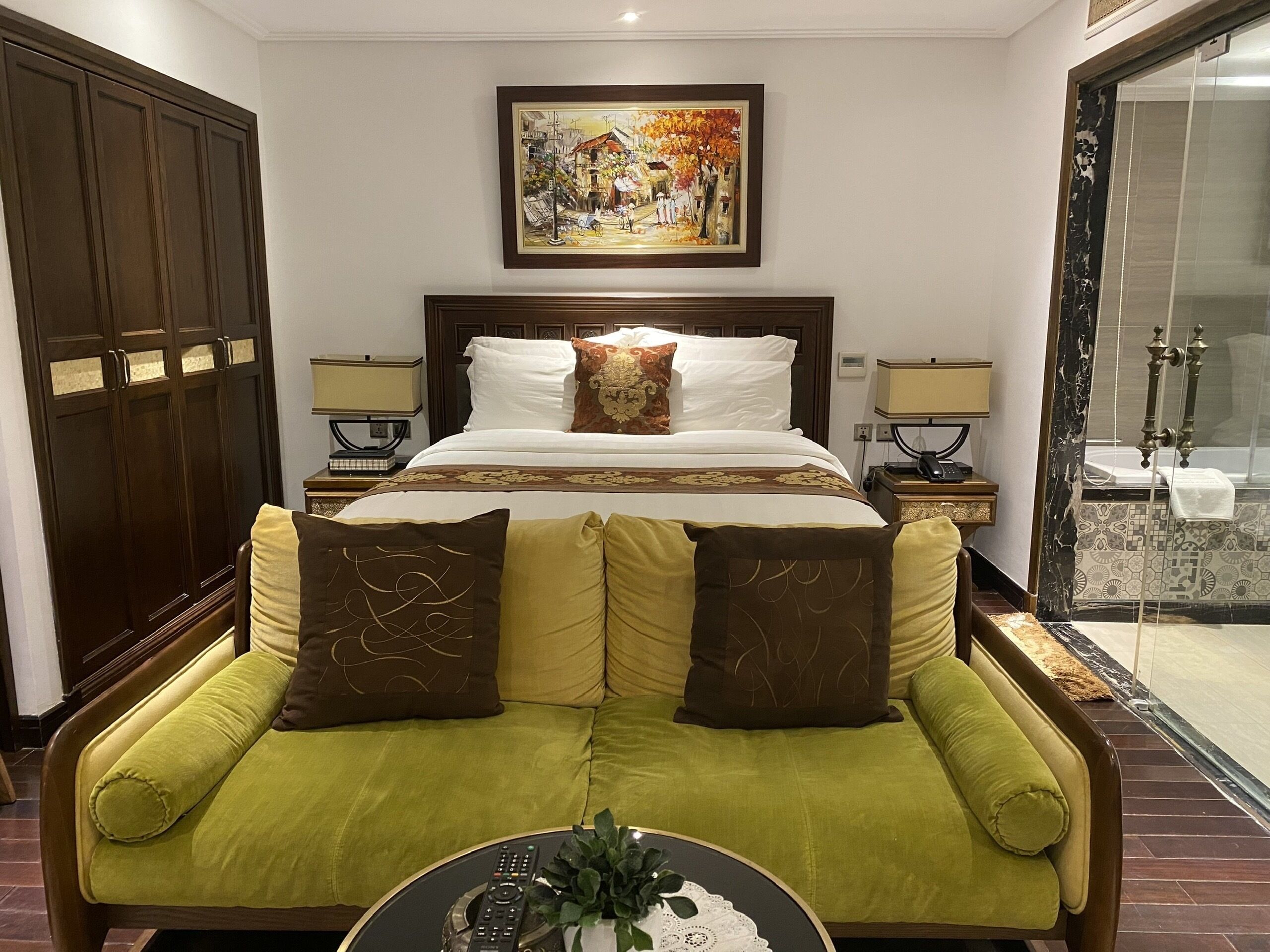 Une chambre d’hôtel avec un grand lit, deux tables de chevet avec des lampes, un canapé vert, un tableau au mur et une salle de bain avec une porte en verre.