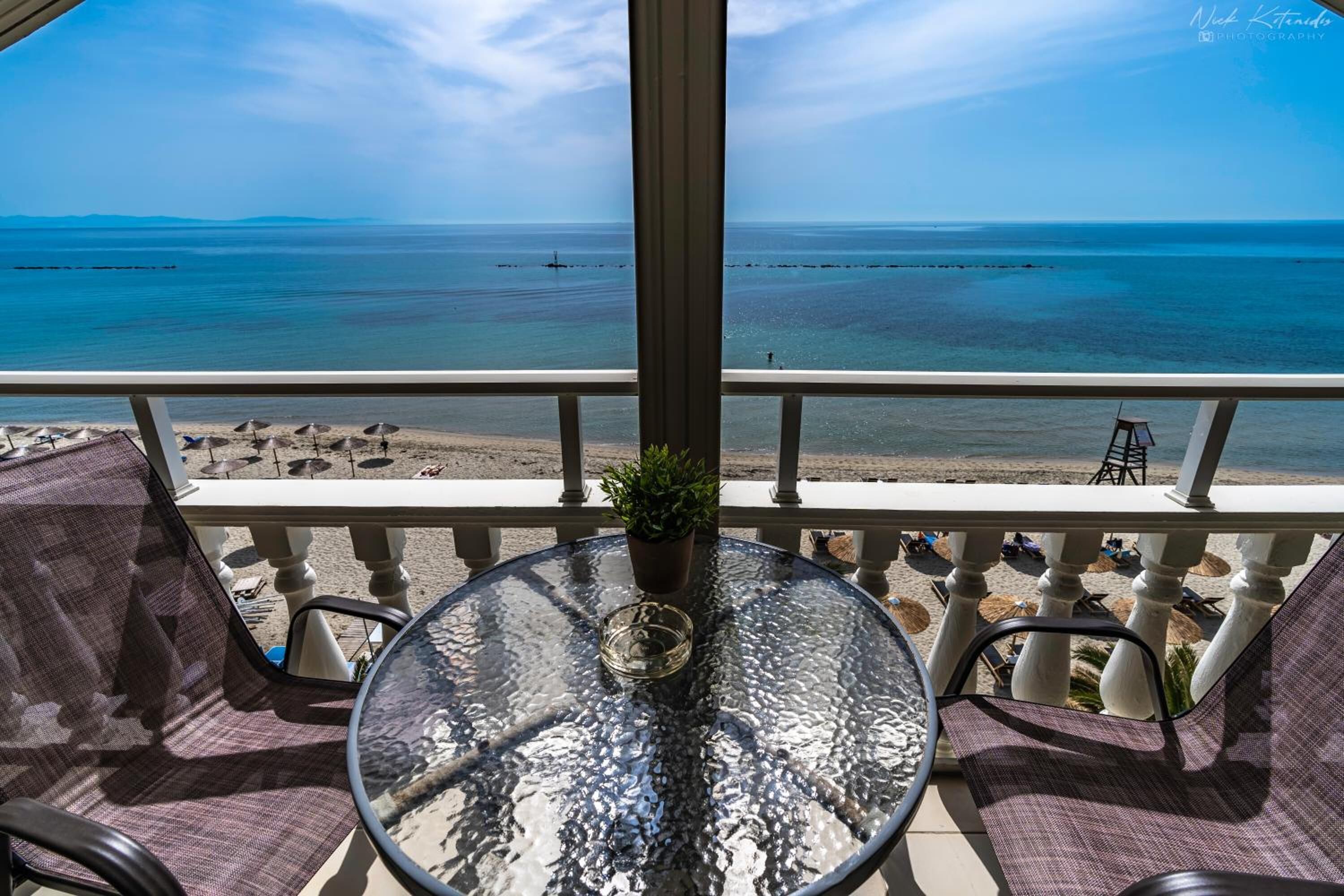 Un balcón con una mesa de vidrio, sillas y una planta en maceta. Se ve una playa y el mar.