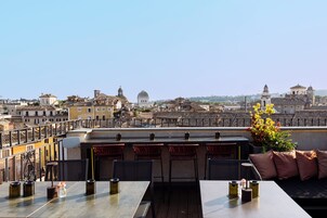 A rooftop terrace with a dining area, chairs, and tables, overlooking a cityscape with historic buildings.