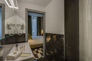 A modern hotel room with a black and white checkered floor, a bed with a green curtain, and a bathroom with a mirror, hairdryer, and toiletries.
