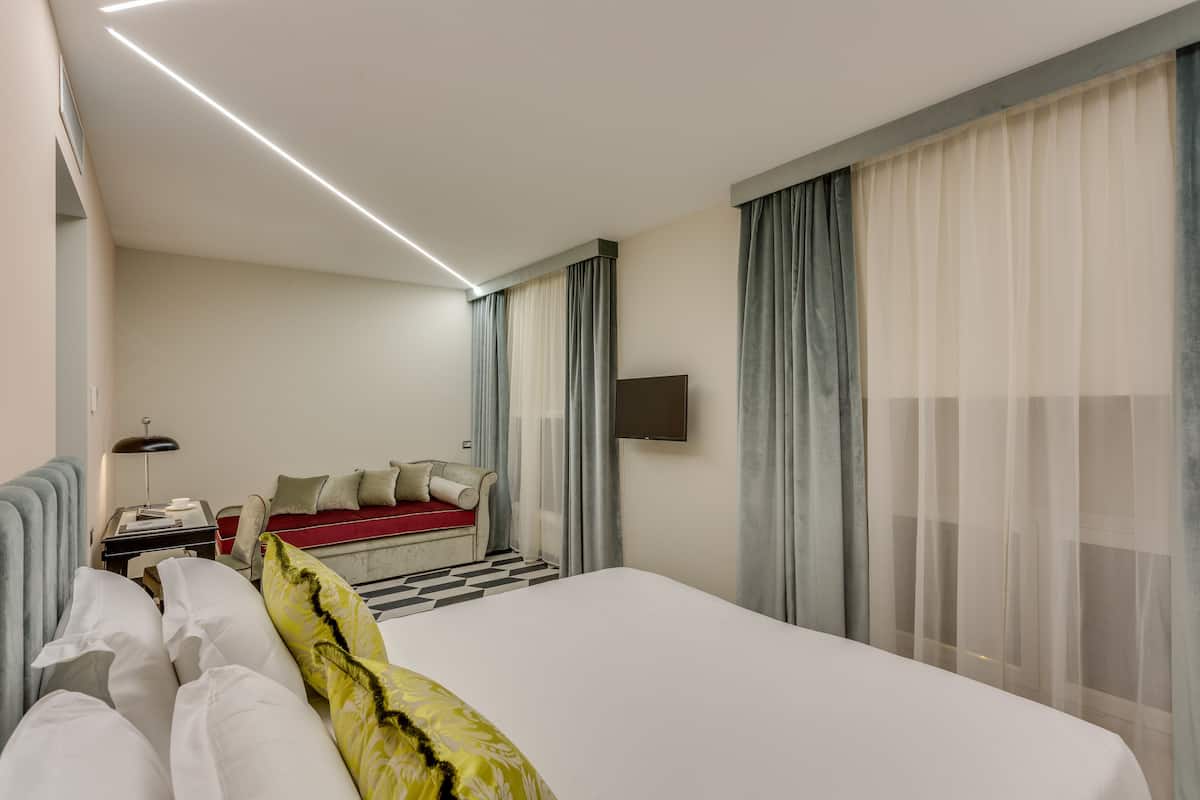 A modern hotel room with a large bed, a desk with a lamp, a TV, and a seating area with a red sofa and cushions.