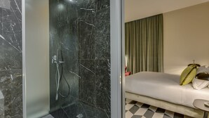 A modern bathroom with a glass-enclosed shower and a white sofa with a green pillow in the adjacent room. - Otivm Hotel (Rome)