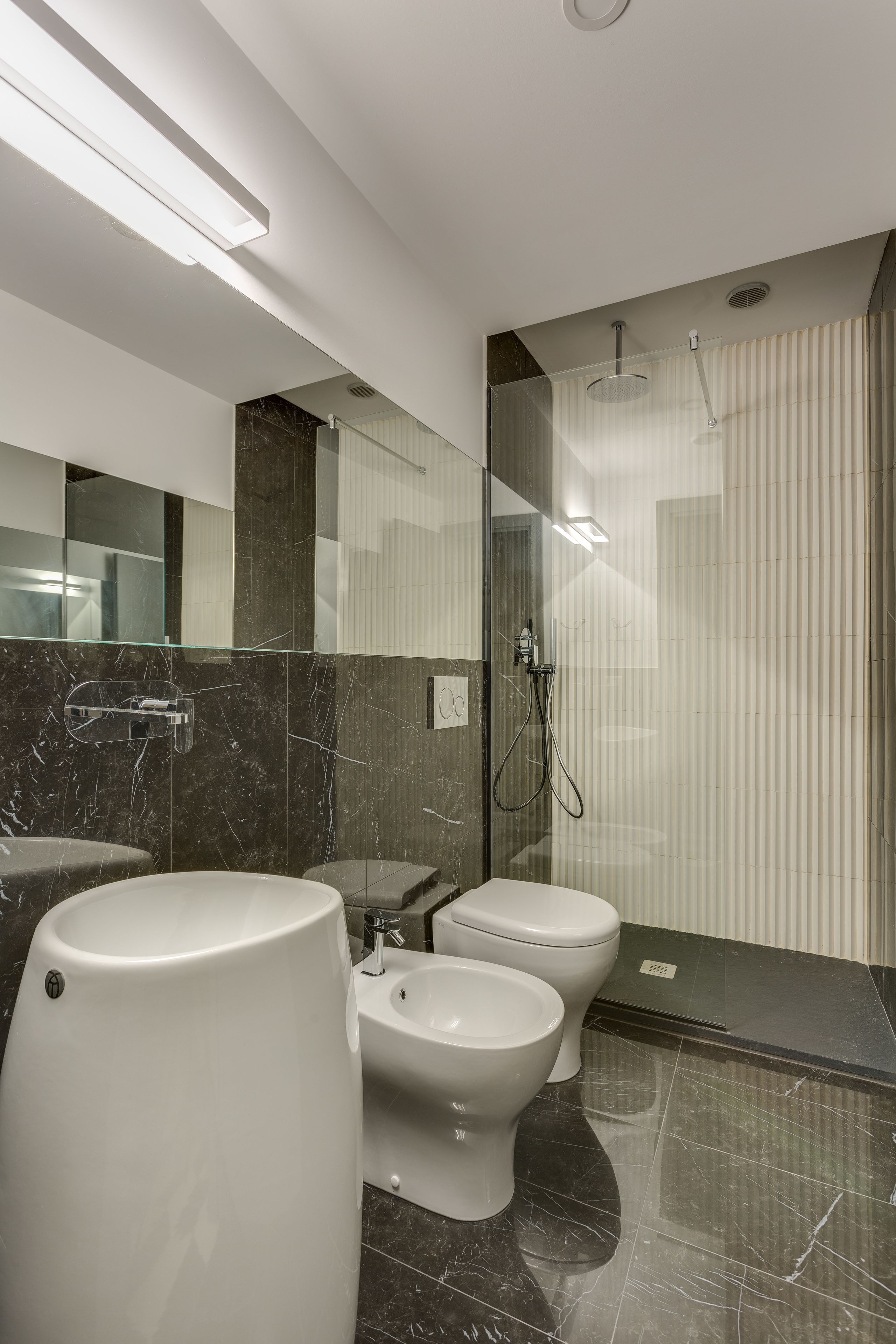 Een moderne badkamer met een marmeren wastafel, twee toiletten, een bidet en een douchecabine met glazen wanden.