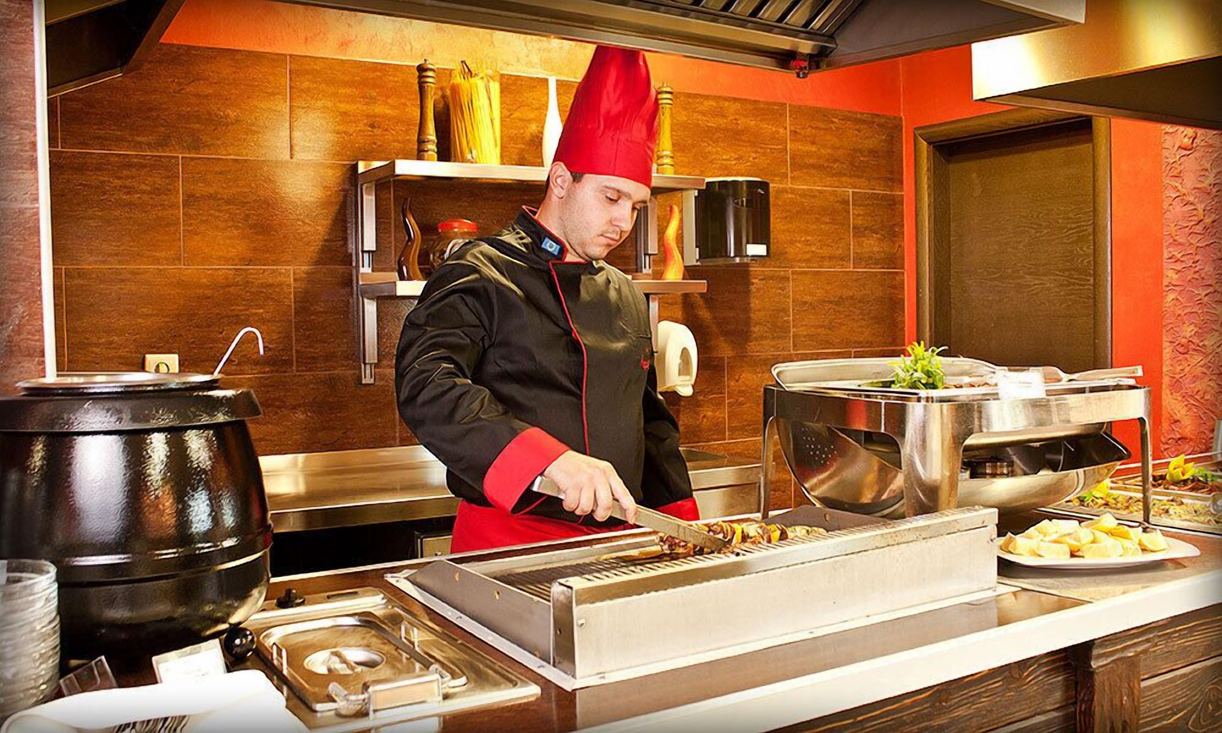 A chef in a red hat preparing food in a kitchen with a buffet setup.