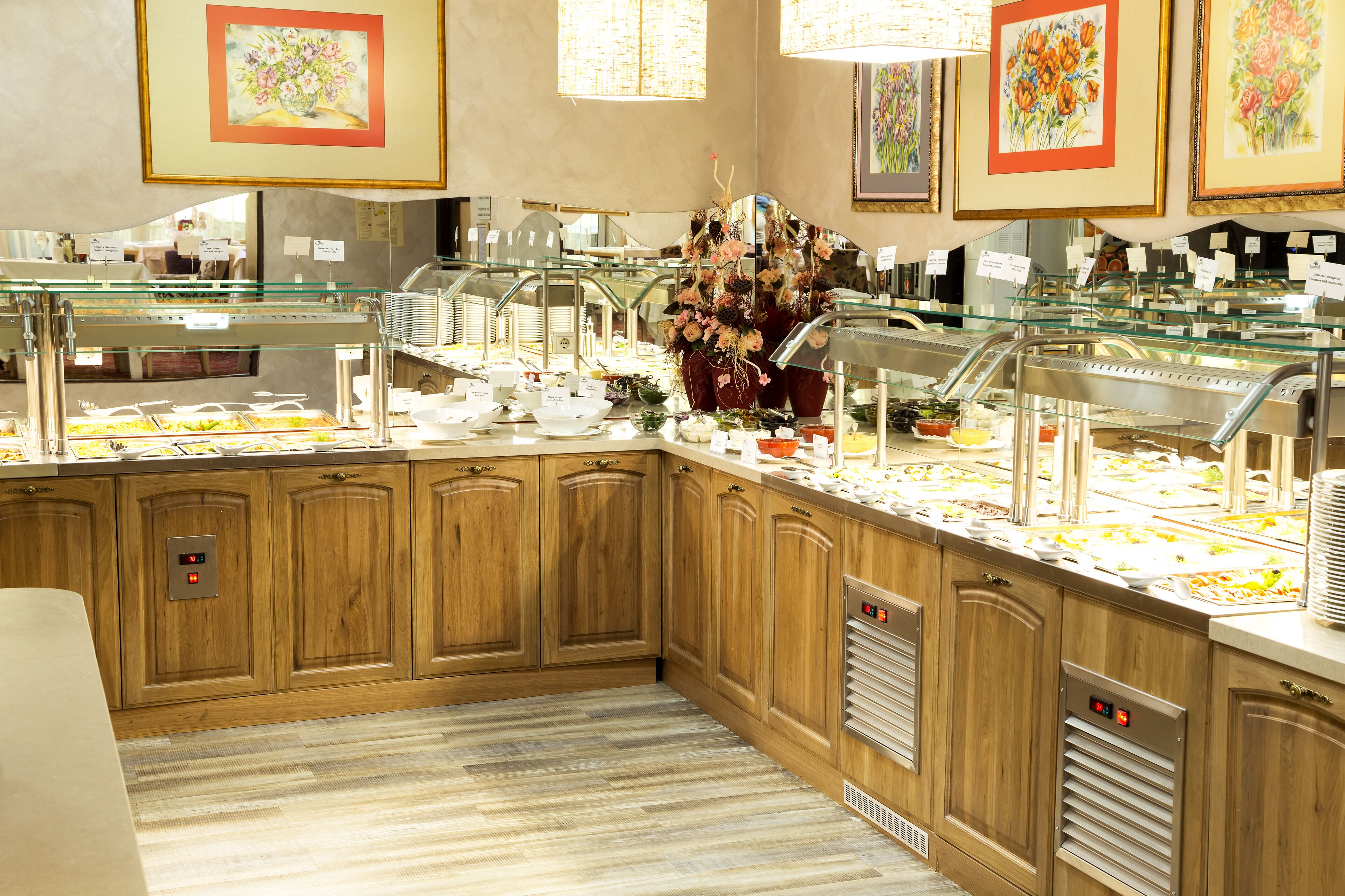 Uma área de buffet com várias estações de comida, armários de madeira e quadros com flores nas paredes.