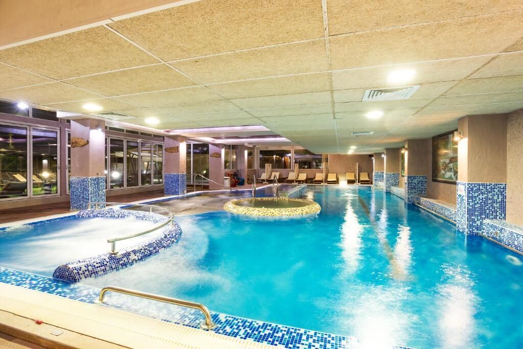 A large indoor swimming pool with a tiled edge, a hot tub, and lounge chairs around the pool area.