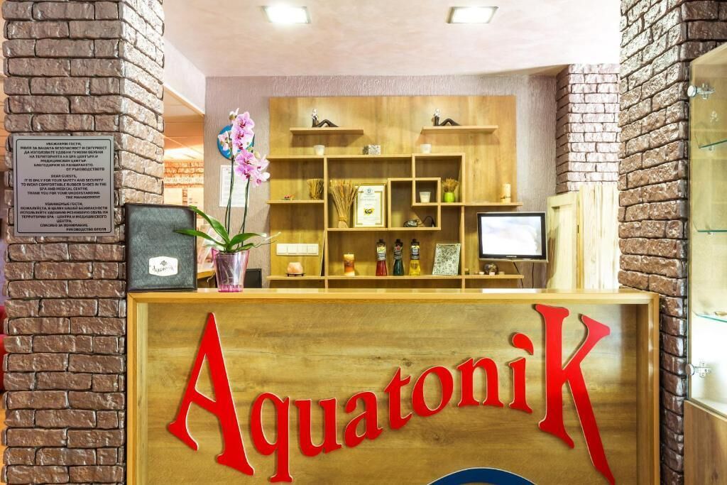 A reception desk with the name 'AquatoniK' in bold letters, wooden shelves with various items, a potted orchid, and a computer monitor.