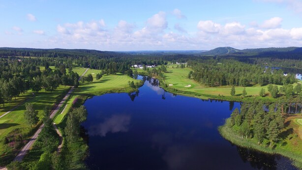 Un campo de golf con un lago, rodeado de colinas boscosas y un cielo despejado.
