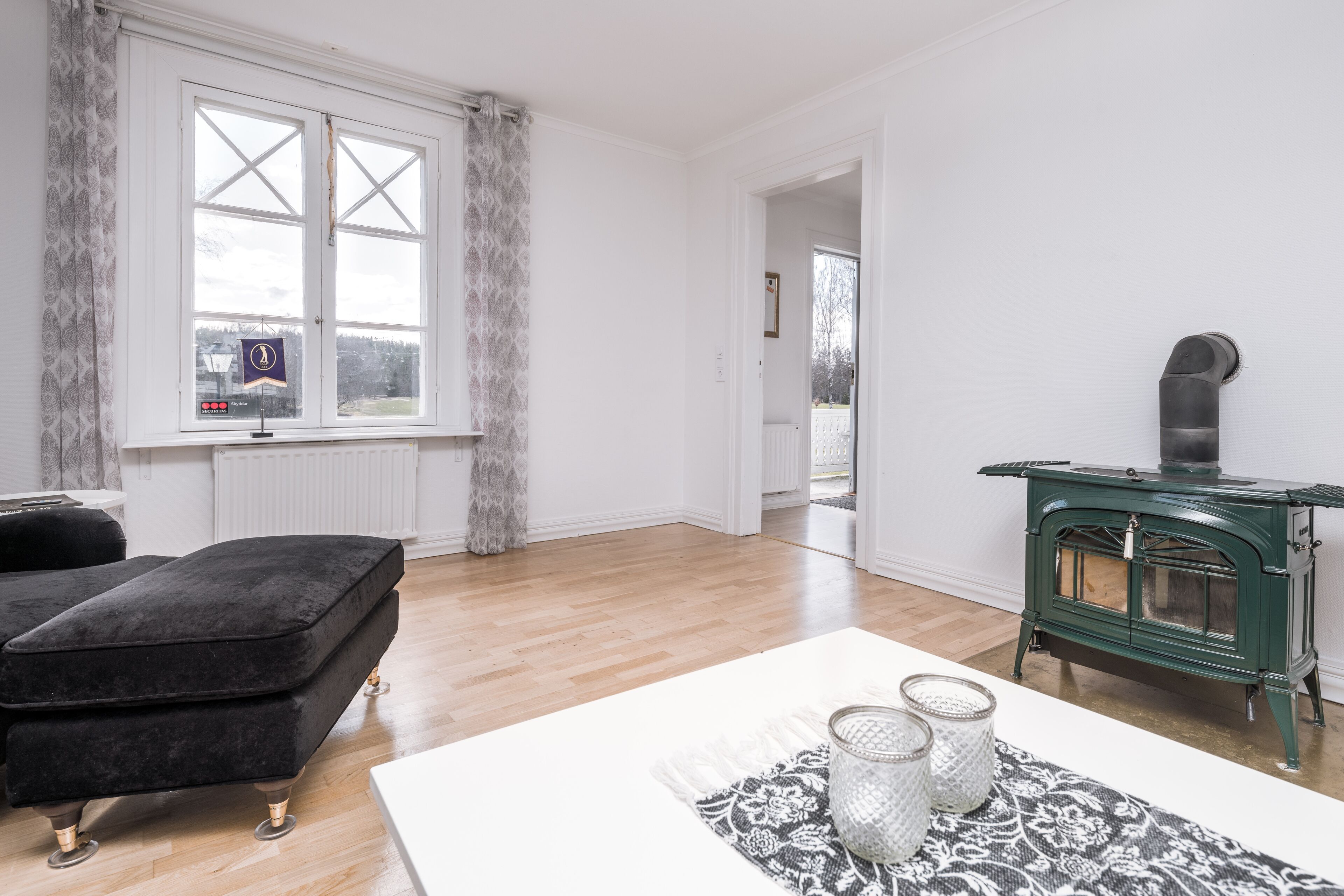 A spacious room with a large window, a green stove, and a black ottoman.