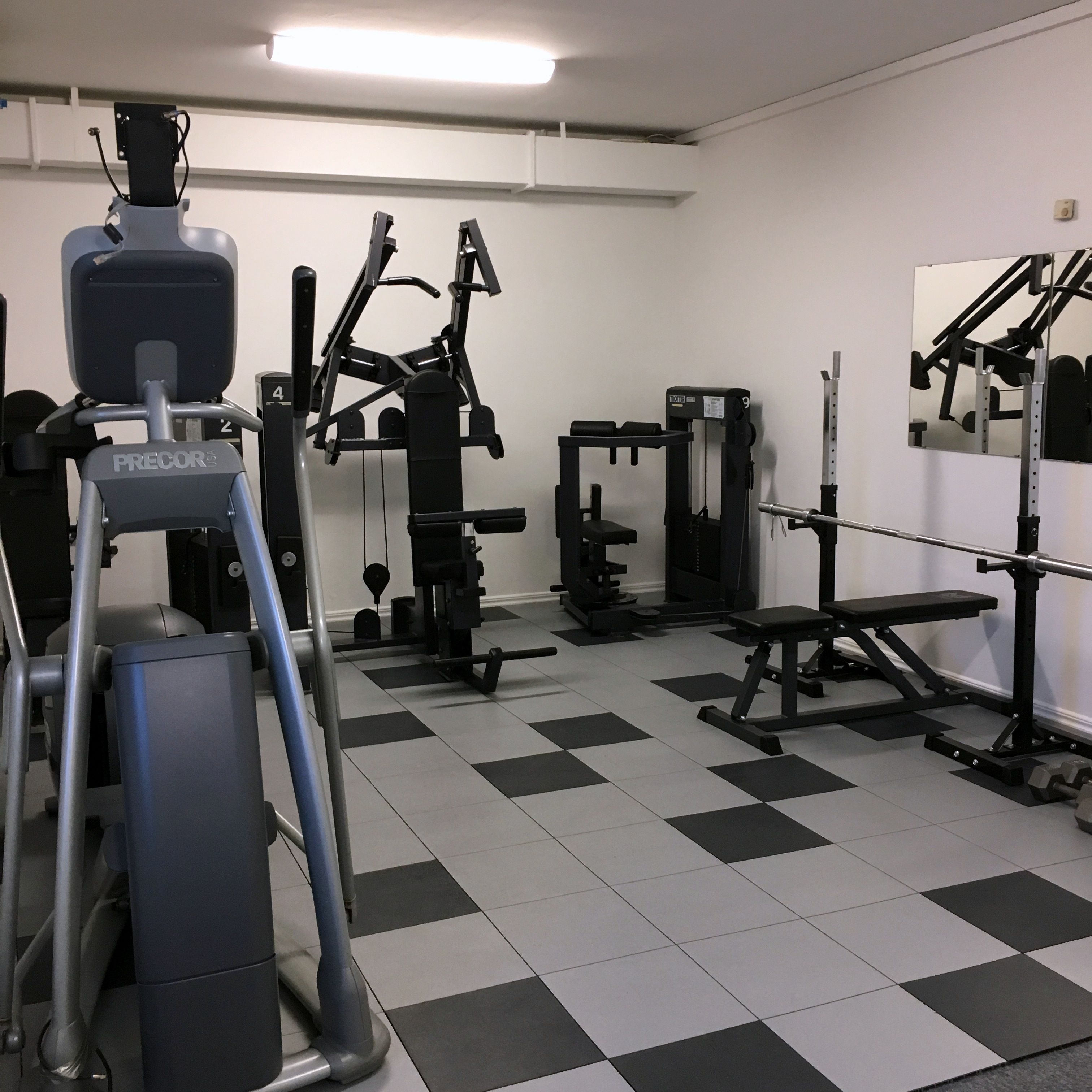 Een sportschool met diverse fitnessapparaten, waaronder een PRECOR loopband, krachtapparaten en een bankje.