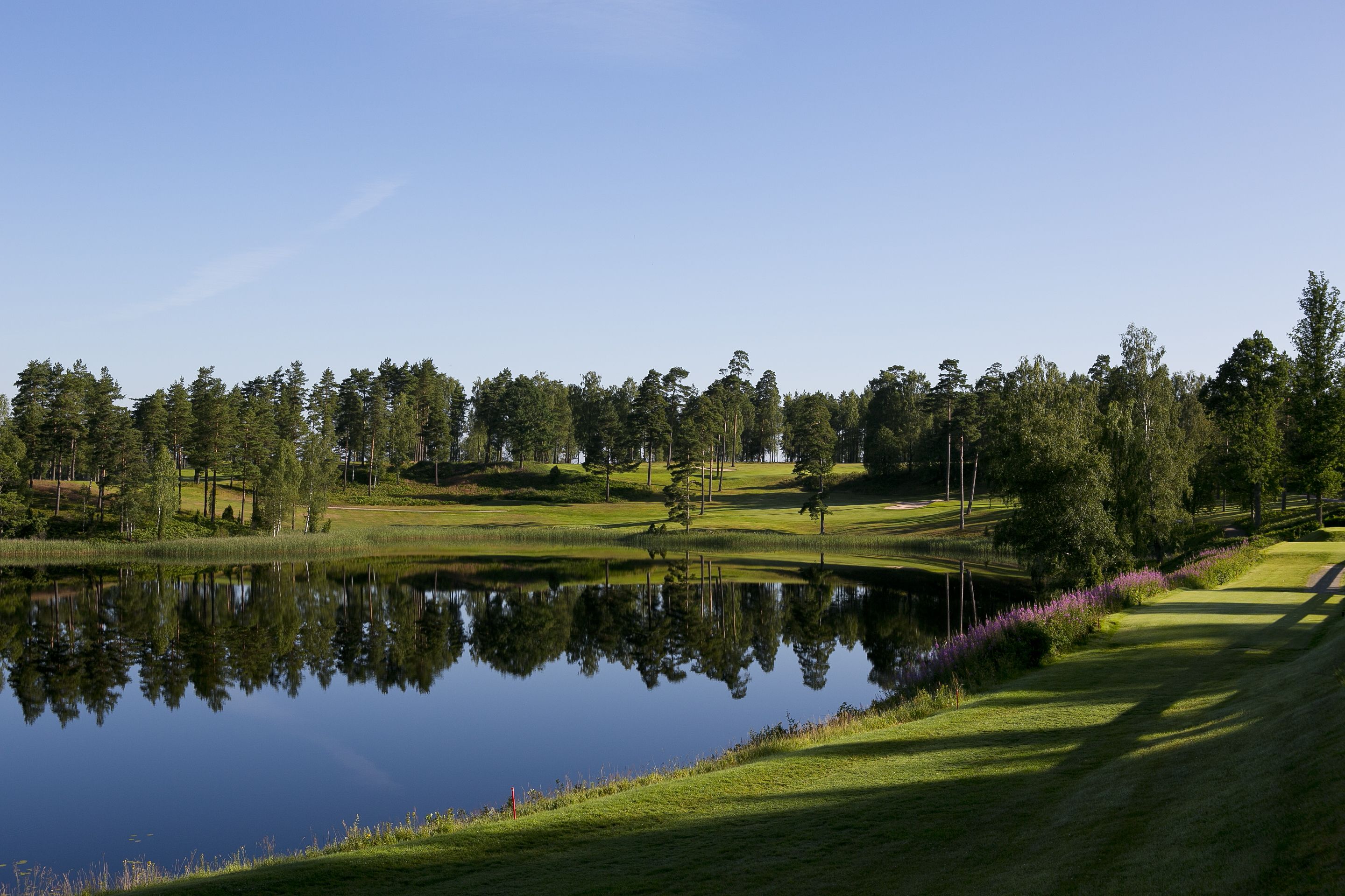 Un campo de golf tranquilo con un estanque que refleja árboles y un cielo despejado.