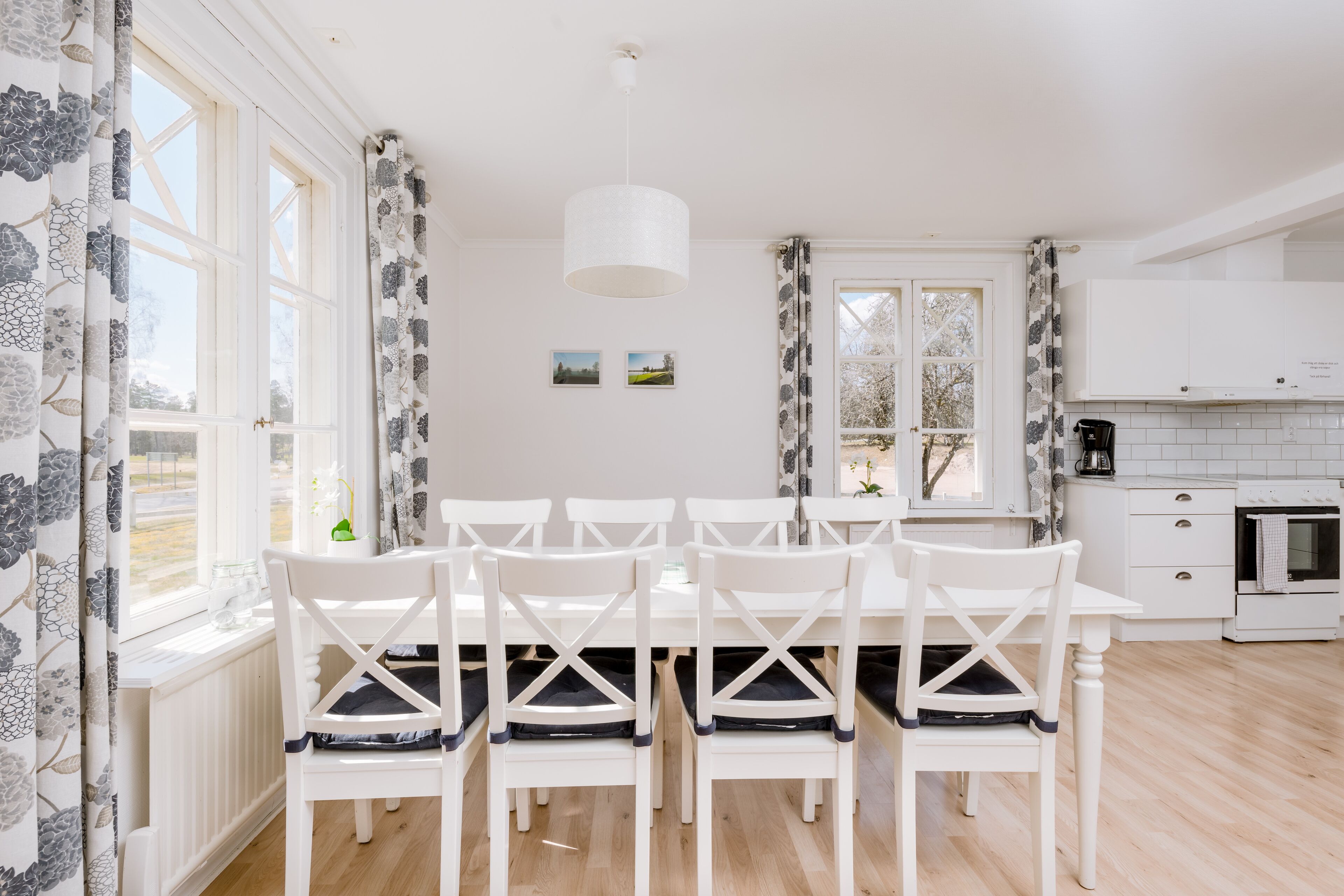 A dining area with a white table and chairs, a hanging light fixture, and a view of the outdoors through large windows.