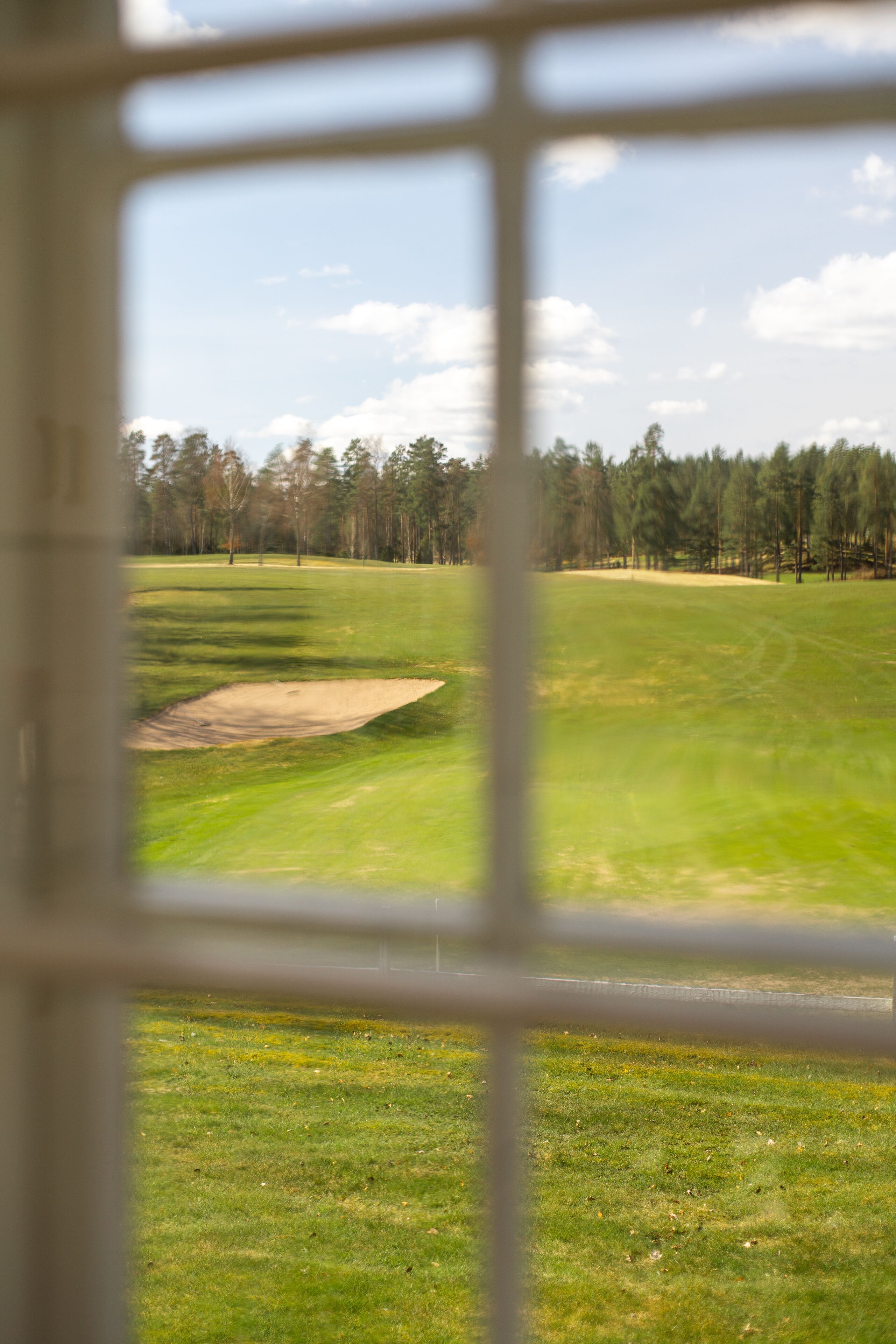 Blick durch ein Fenster auf einen Golfplatz mit einem Bunker und einer Baumreihe im Hintergrund.