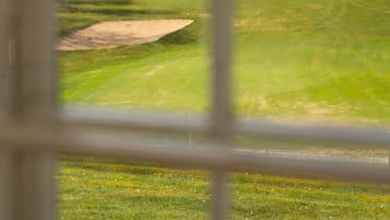 Blick durch ein Fenster auf einen Golfplatz mit einem Bunker und einer Baumreihe im Hintergrund.