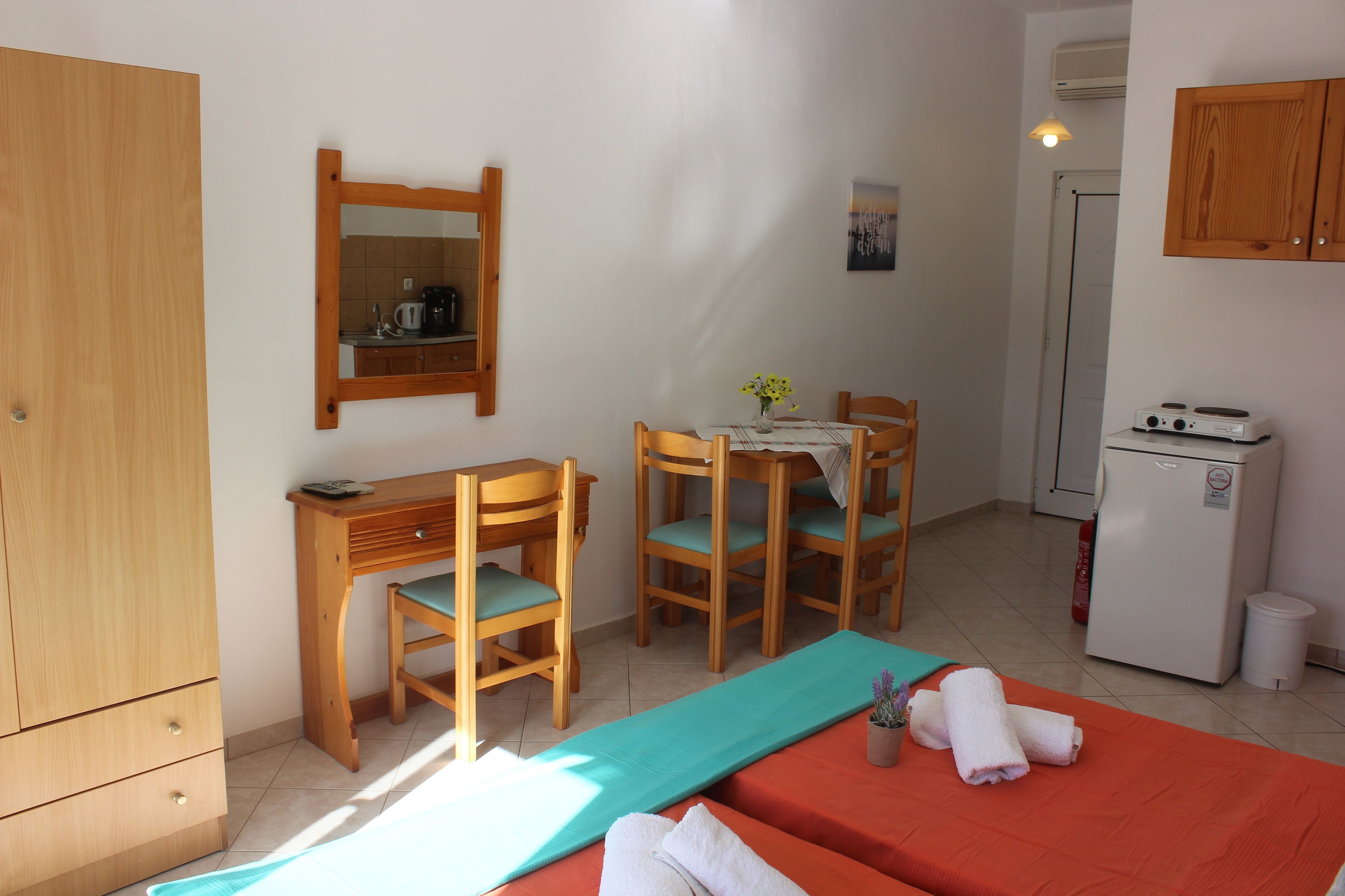 Une chambre à coucher comprenant un lit, un bureau, deux chaises, une petite table avec un vase, un miroir, une armoire, un mini-réfrigérateur et un meuble de rangement.