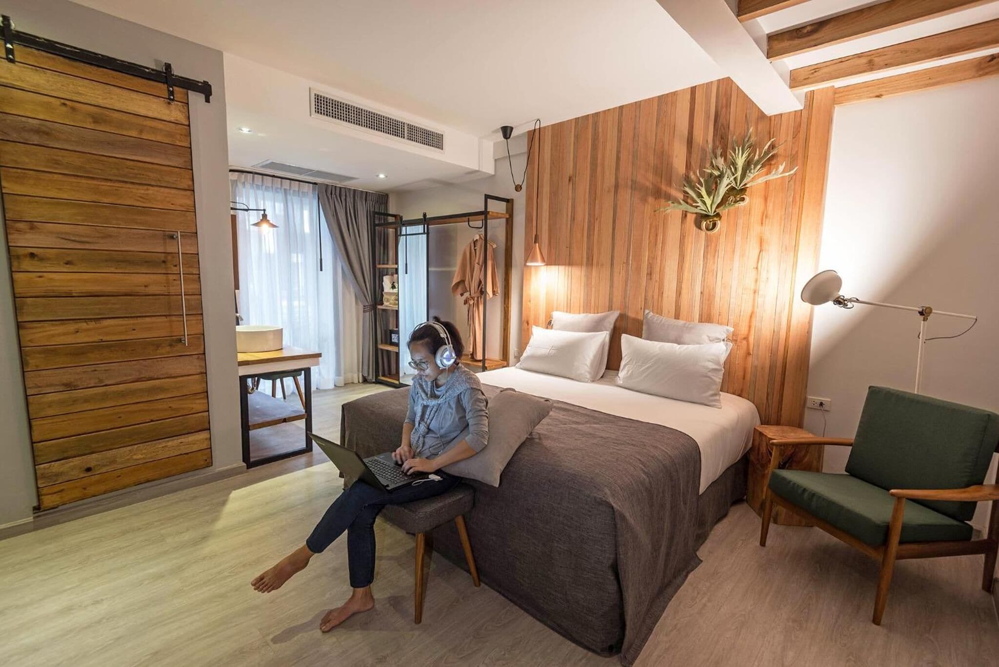 A modern bedroom with a large bed, a wooden wall panel, a green chair, and a person using a laptop.