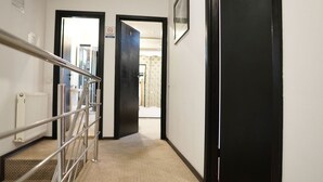 Un pasillo con barandilla de metal, un radiador y suelo cubierto de alfombra, que conduce a una puerta.
