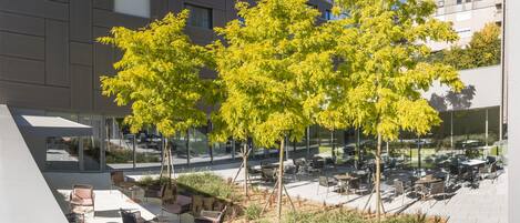 An outdoor seating area with tables and chairs, surrounded by trees and a building in the background.